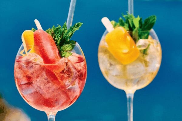 Colorful summer spritz cocktails with fruit popsicles and mint, served over ice in wine glasses at Bites & Bubbles by Sunsets with Aegean Sea backdrop