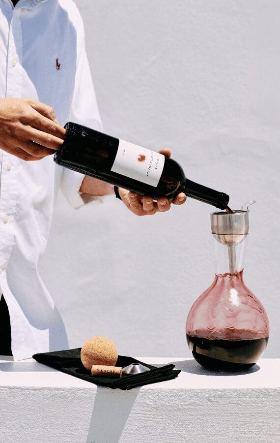 Sommelier decanting a bottle of Santorini red wine into a glass carafe against a minimalist white backdrop at Sunsets Terra