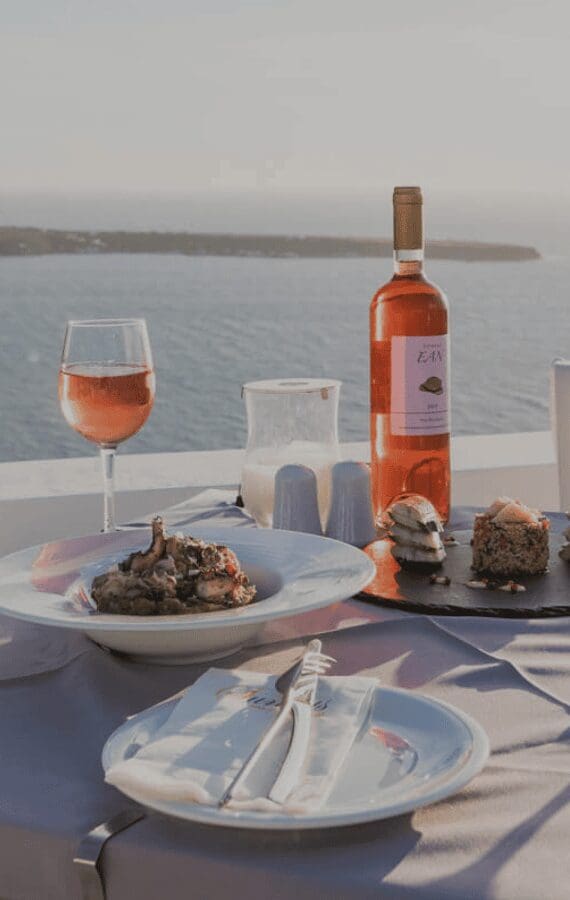 Romantic caldera-side table at Santorini Sunsets with rosé wine, gourmet main course, and desserts overlooking the Aegean at golden hour