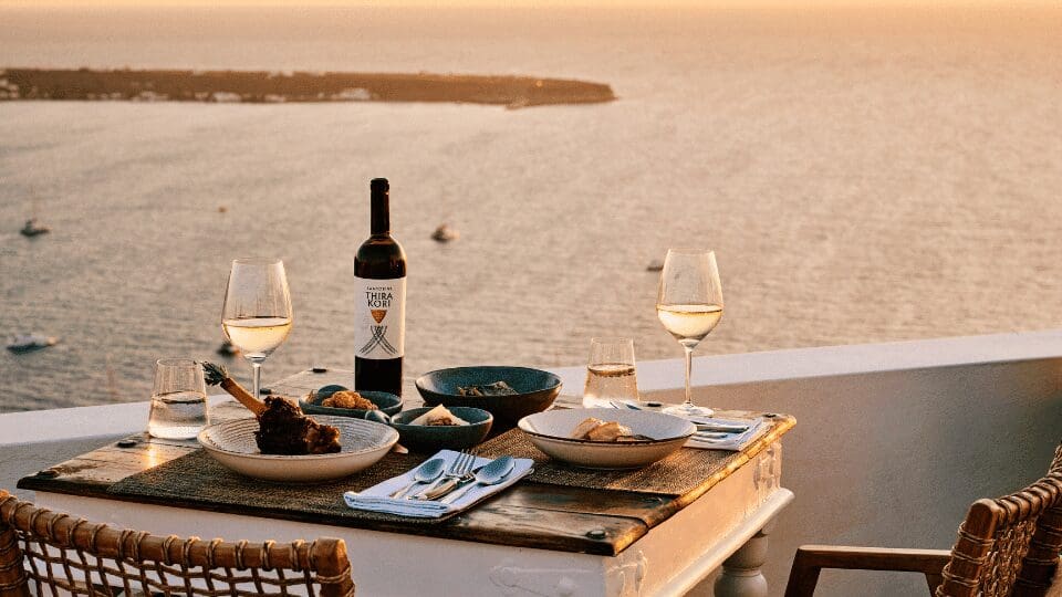 Cliffside sunset dining setup in Oia with glasses of white wine, local dishes, and a bottle of Santorini wine overlooking the Aegean Sea and caldera views