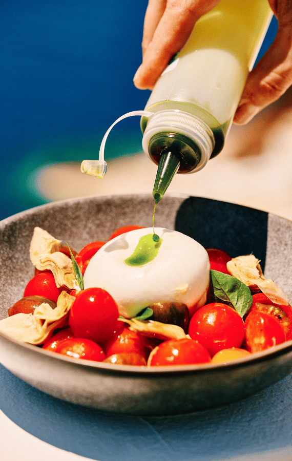 Fresh burrata salad with cherry tomatoes, basil, and artichokes being drizzled with herb oil, served at Santorini Sunsets with a sea view backdrop