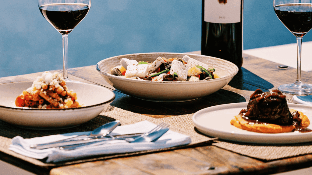 Three Mediterranean dishes—Greek salad, pasta with feta, and slow-cooked beef—paired with red wine, served on a rustic table overlooking the Aegean in Santorini