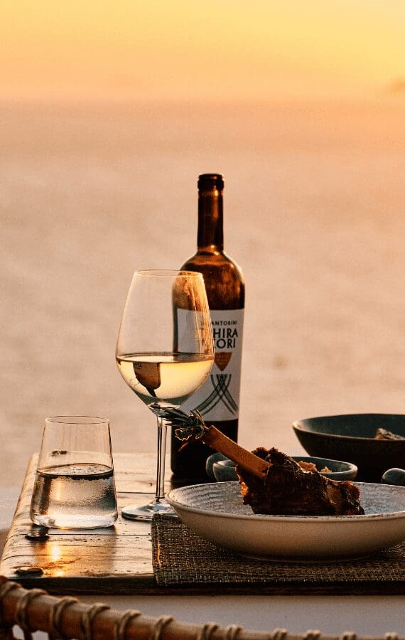 Glass of white wine and slow-cooked lamb shank served at sunset on a cliffside table in Santorini, with golden light reflecting off the Aegean Sea
