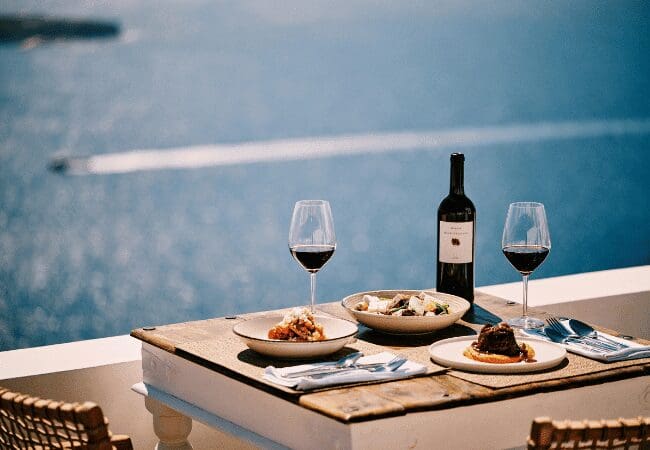 Seafront table at Sunsets Santorini with Mediterranean dishes, red wine, and panoramic Aegean views