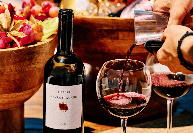 Pouring Santorini-grown Sigalas Mavrotragano red wine into glasses beside a fruit bowl at Bites & Bubbles by Sunsets