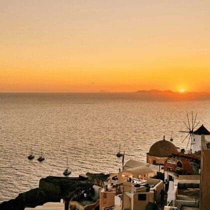 Sunset over the Aegean Sea with sailboats and Oia’s windmill in view near Sunsets Santorini