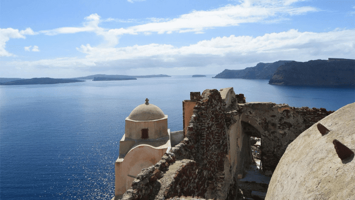 Panoramic view from Oia Castle ruins in Santorini with domed chapel and caldera cliffs overlooking the Aegean Sea before sunset hour
