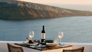 Sea-view terrace table with Greek dishes, a wine bottle and two white-wine glasses overlooking the caldera in Oia, Santorini.