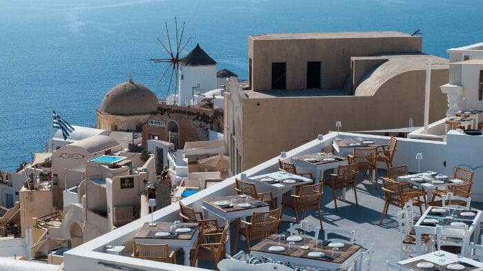 Cliffside Santorini Sunsets restaurant terrace in Oia with caldera view, set with tables overlooking Santorini’s windmill and blue Aegean Sea.
