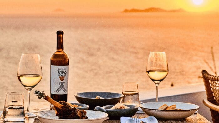 Thira Kori wine bottle and glasses of white wine on a sunset terrace overlooking the Aegean Sea in Santorini.
