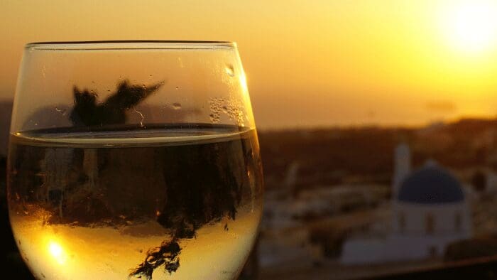 Glass of white wine at sunset with view of blue-domed church and Aegean Sea in Santorini.