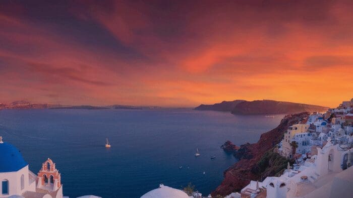 Vivid pink and orange sunset with cotton candy clouds over Oia’s blue-domed churches and caldera in Santorini.