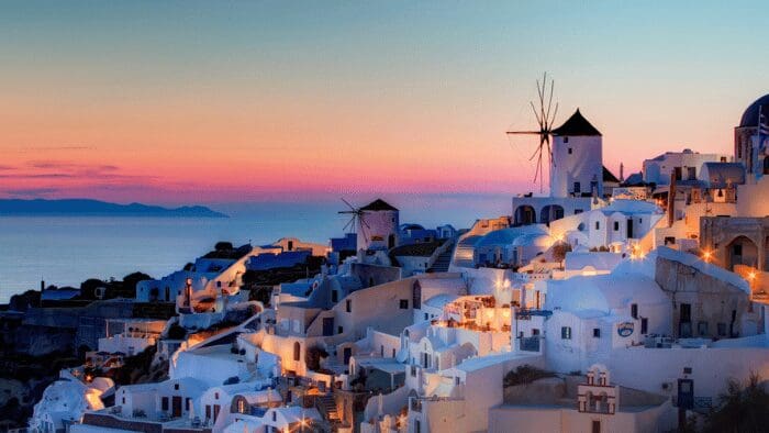 Twilight view of Oia, Santorini with soft purple sky after sunset and illuminated whitewashed houses above the caldera.