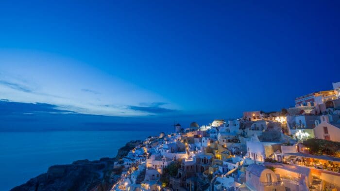 Blue hour panoramic view of Oia, Santorini, Greece, with illuminated whitewashed houses and calm Aegean sea after sunset.