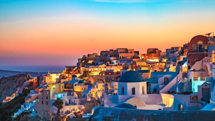 Oia village at dusk in Santorini, Greece, with glowing lights, blue domes, and pastel sunset sky over the caldera.