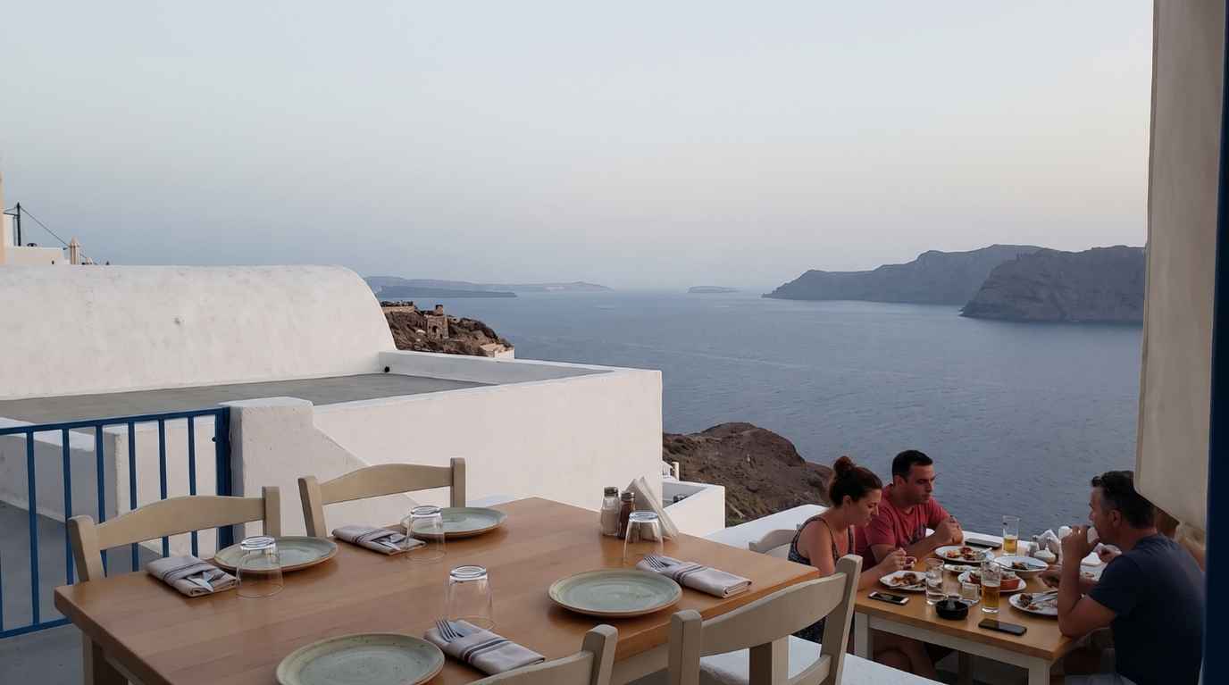 Restaurant table in Oia set for dinner with a neighbouring terrace and other diners between it and the caldera edge